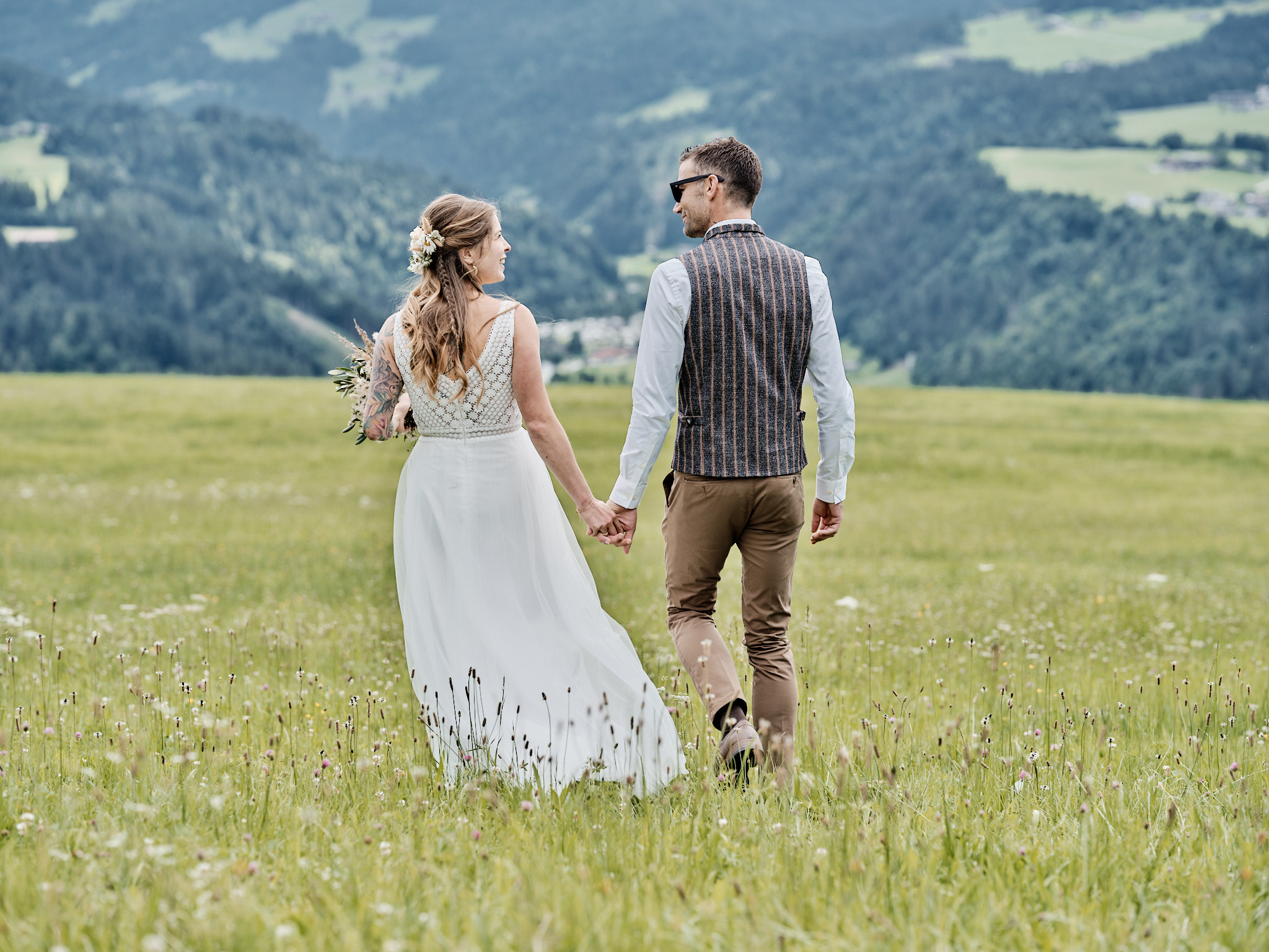 Das Brautpaar spaziert beim Portraitshooting händehaltend durch eine Sommerwiese.