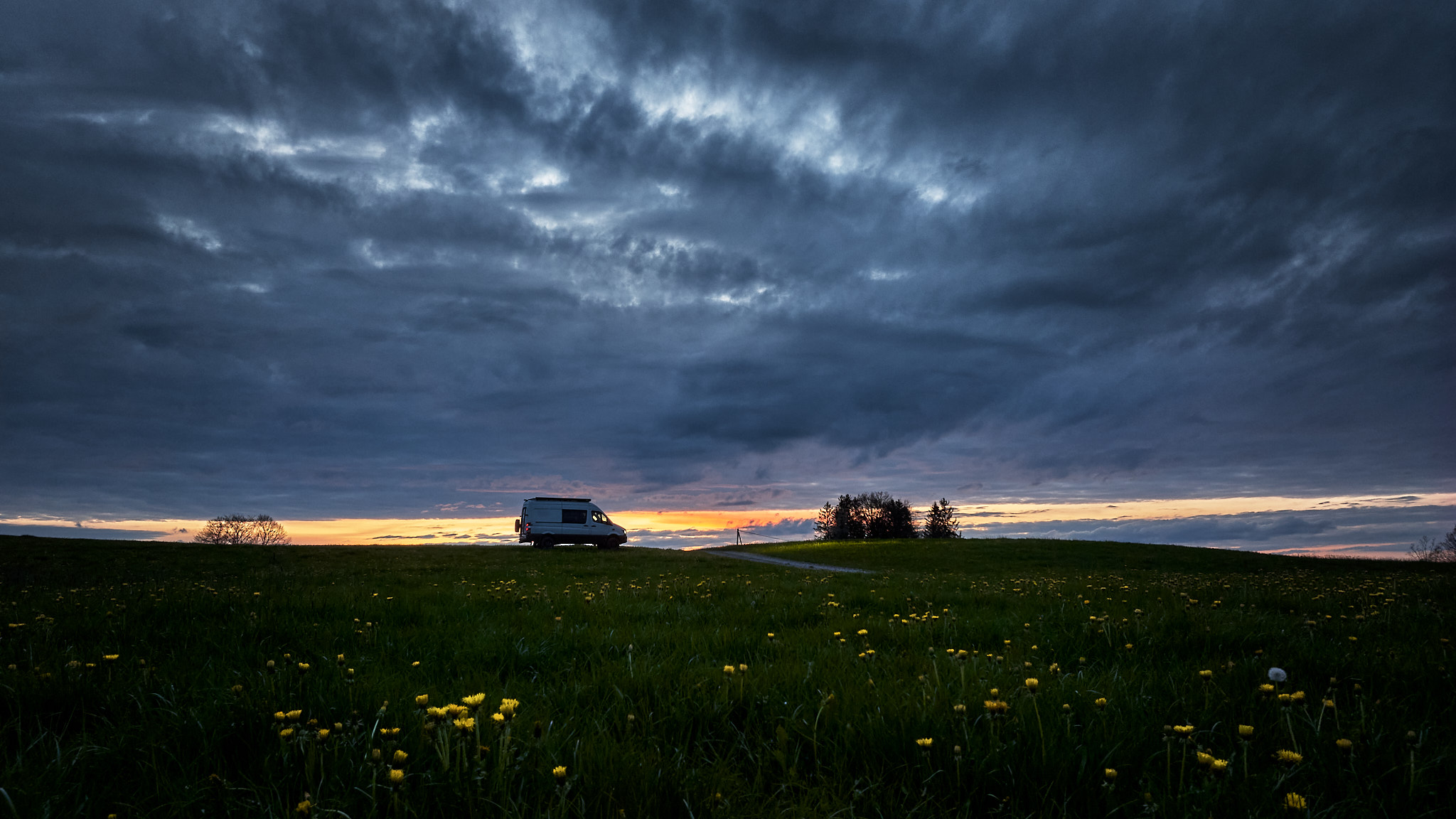 Reise- und Naturfotograf steht mit Camper am Wegrand vor einem Sonnenuntergang unter wolkenverhangenem Himmel. Die Wolken am Horizont leuchten in rötlichen Farben.